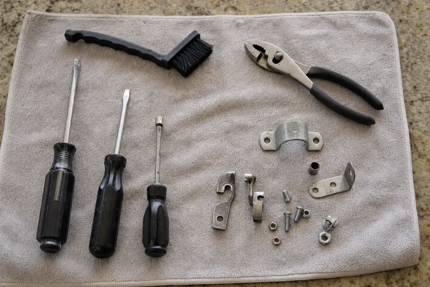 Screwdrivers, a parts brush, slip-joint pliers, a pipe clamp and small brackets and screws laid out on a cloth over a granite worktop