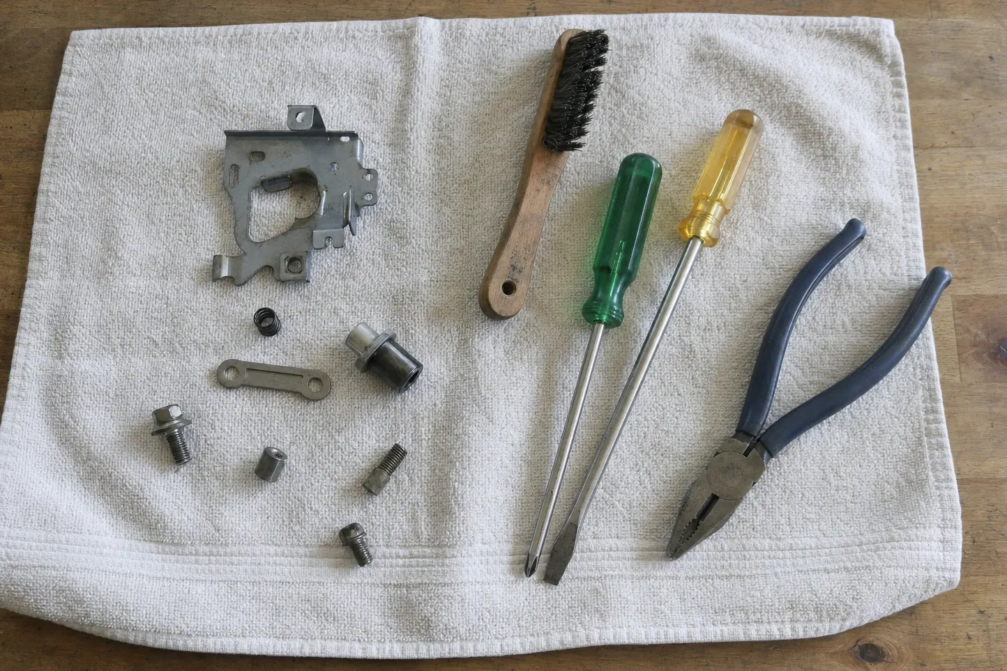 Large stamped mounting plate, coil spring, link bar and bolts laid out on a cloth with a wire brush, two screwdrivers and pliers